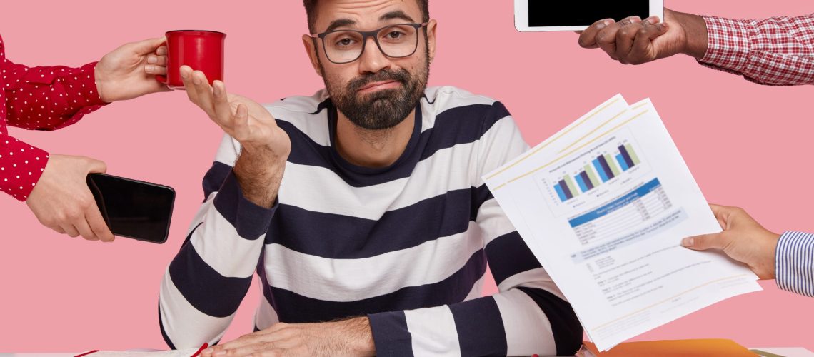 Sleepy inexperienced bearded young man with hesitant expression, spreads hand, wears spectacles and striped sweater, sits at desktop with organizer and paper documents, isolated on pink wall